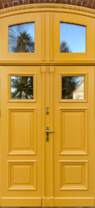 Dwuskrzydłowe drzwi drewniane z podwójnym ramiakiem z naświetlem łukowym, ozdobione frezowanymi listwami i opaską, w ceglanej elewacji